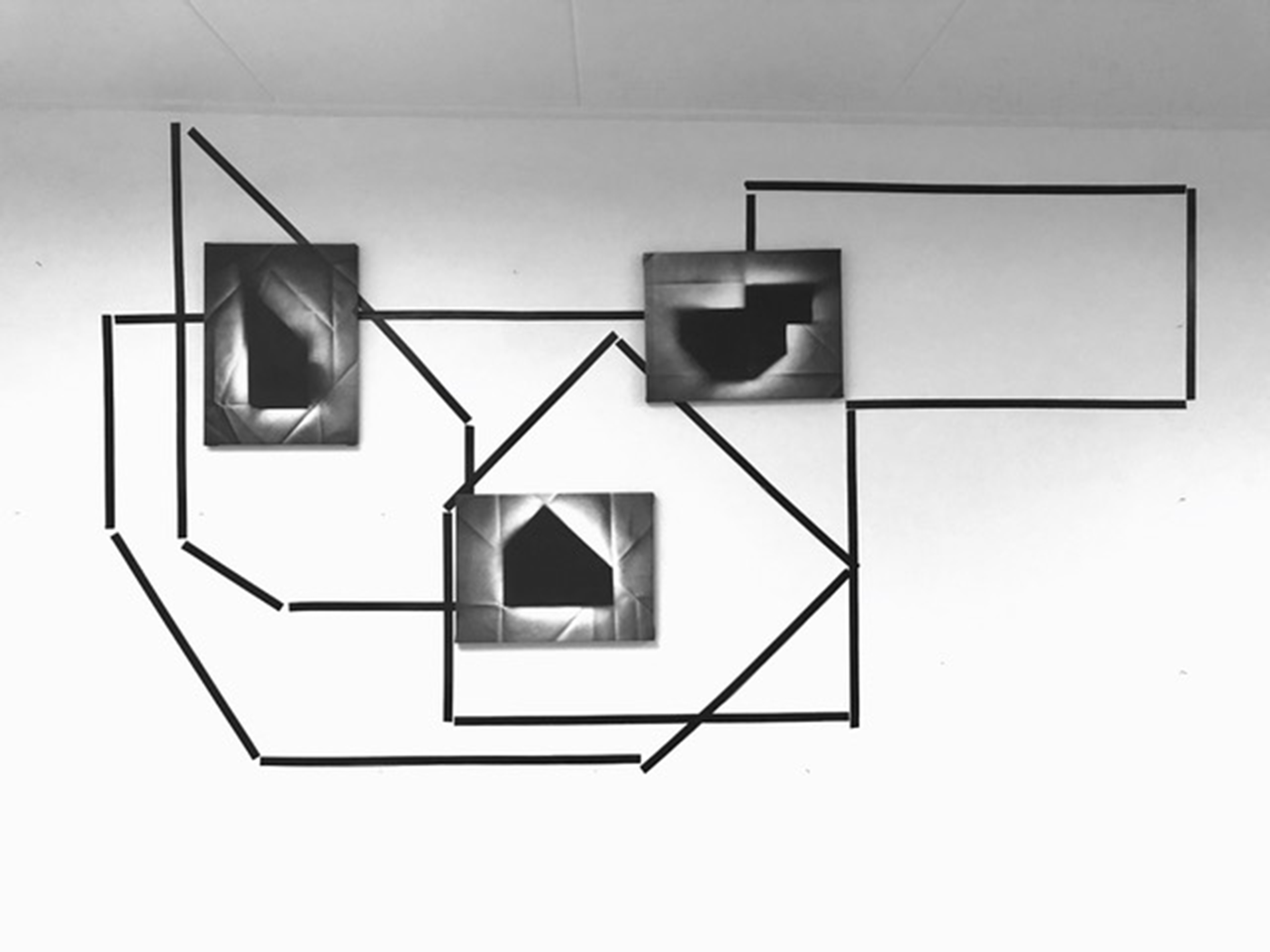 drie geometrische zwart-witte schilderijen hangen op verschillende hoogtes aan de muur over een lijnenspel van zwarte tape